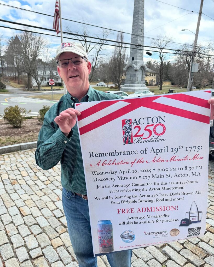 A man in an Acton 250 hat holds a larg poster in front of the monument in Acton Center. The poster advertises the Remembrance of April 19th event at the Discovery Museum (on Wednesday, April 16).