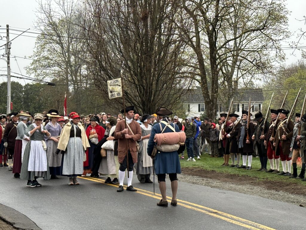 A group of men and women, mostly in colonial garb are lined up on a road. To the side, a group of people in Minuteman dress shoulder long guns.