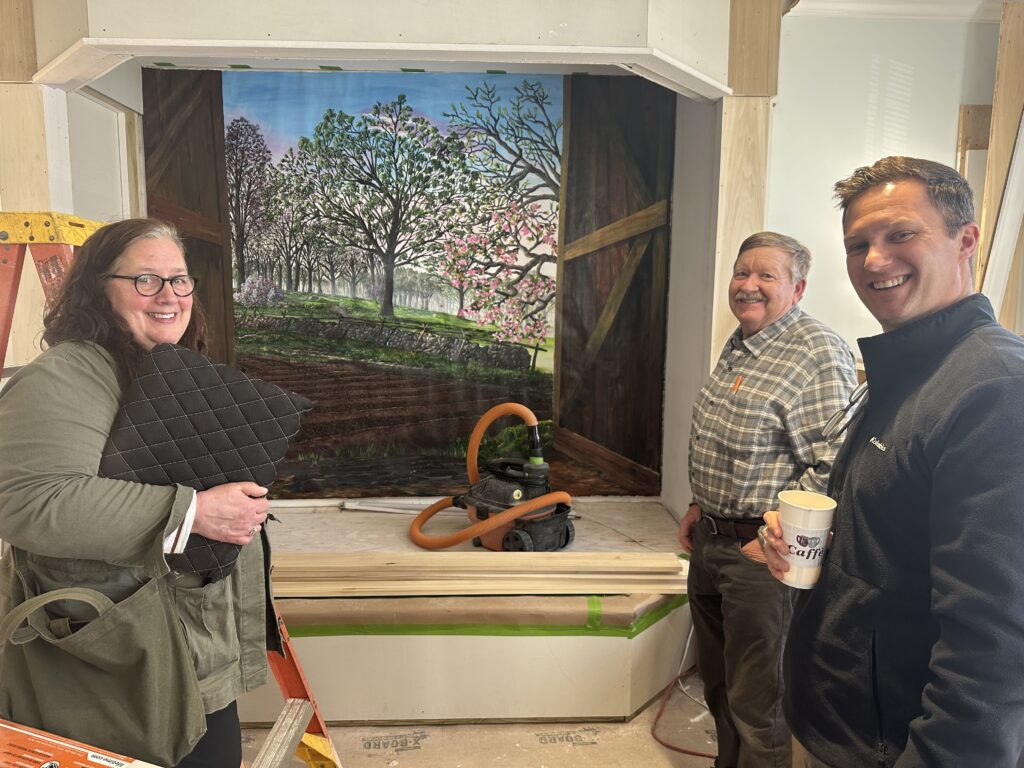 A woman and two men stand in front of a painted backdrop of flowering trees. There's plywood and woodworking tools out, indicated a work in progress.