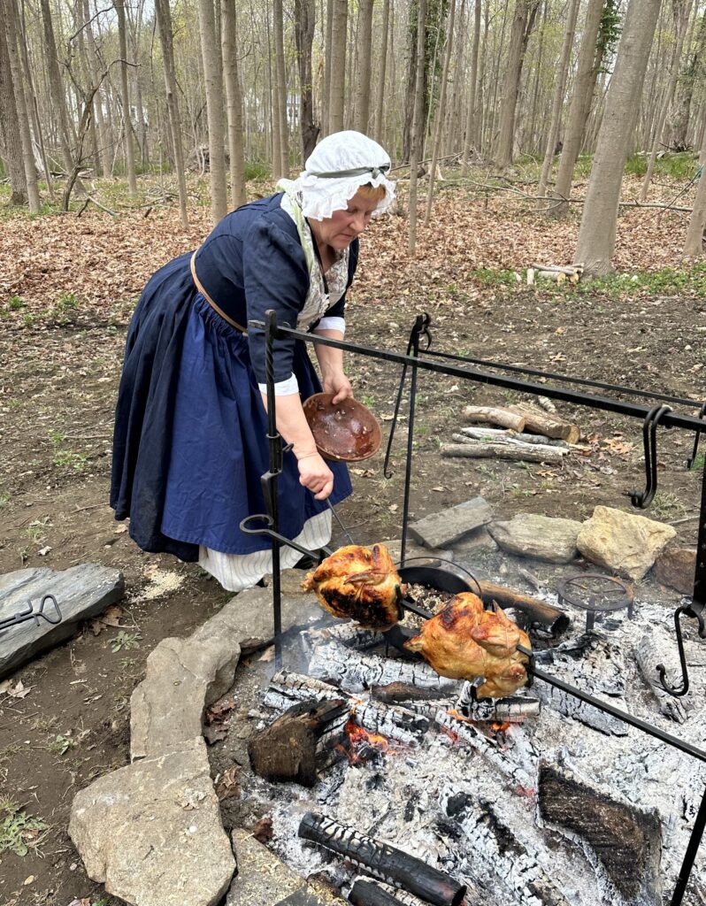 A woman in an 18th Century dress (complete with mob cap) turns two chickens on a spit over a wood fire.