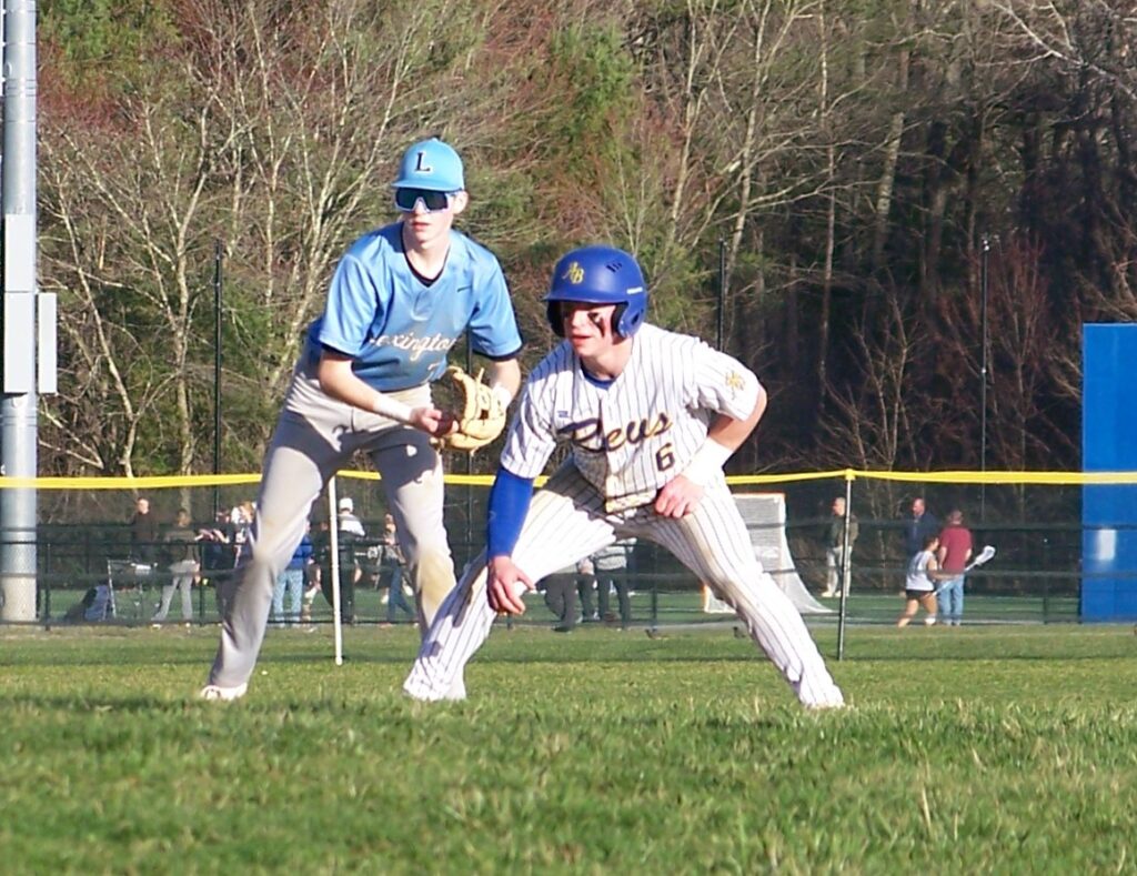 Two baseball players. The player on the left wears a Lexington uniform and is waiting for a ball to come in. The player on the right is on base and looking for a chance to run.