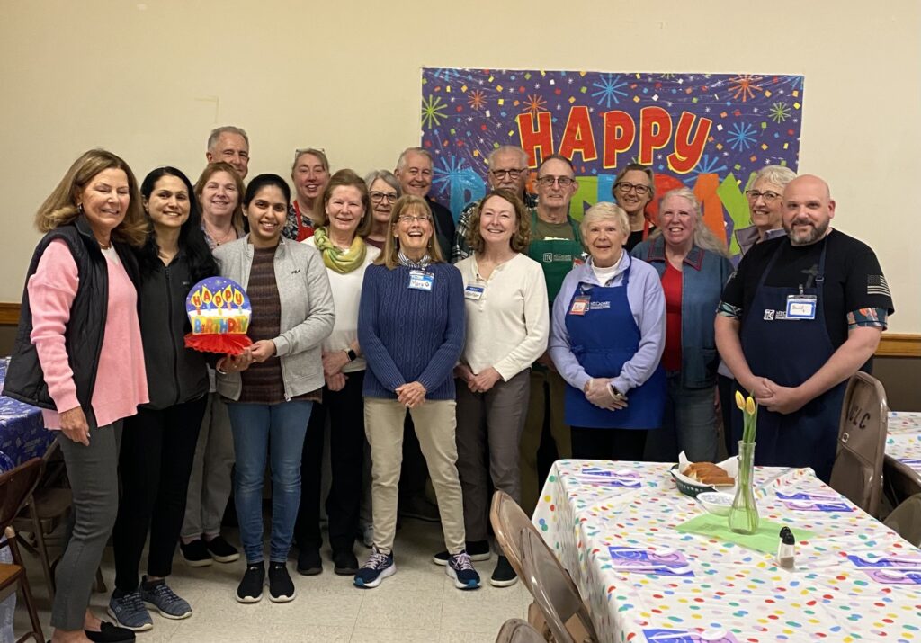 A group of men and women in front of a Happy Birthday sign. In the foreground is a brightly decorated table, ready for guests to come in.
