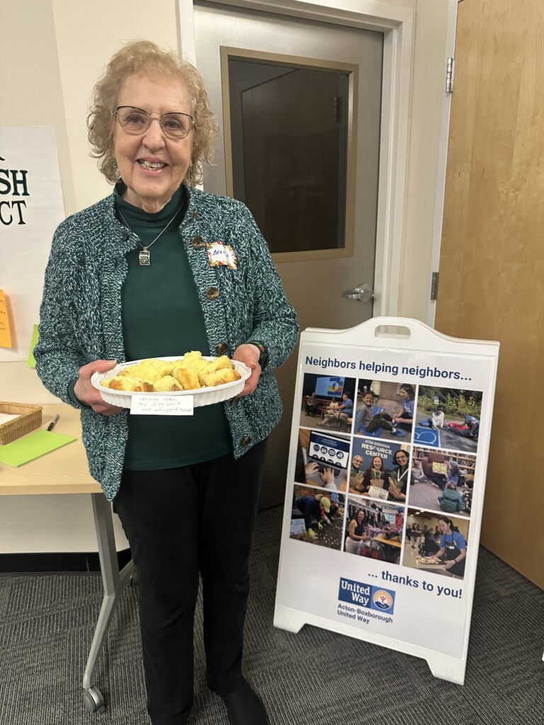 An older woman stands with a platter of cake next to a United Way poster.