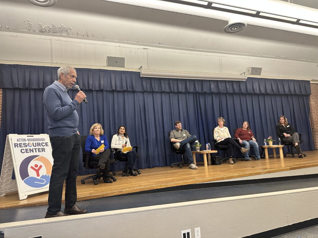 A man in a blue sweater stands and speaks on a stage with several other seated people.